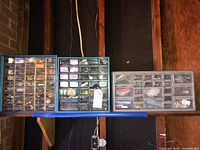 Three plastic storage units for hardware, blue and gray, each with many small compartments and drawers filled with assorted nuts, bolts, screws, nails, washers, and plugs.