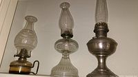 Three vintage oil lamps placed side by side showing different glass bases and chimneys with metal components, varying heights from about 14 to 24 inches.
