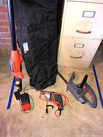 Black & Decker tree trimmer with black collection bag, battery charger, and battery pack visible on the floor