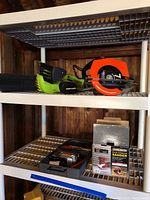 Shelf with green and black Portland electric chainsaw and orange-bladed circular saw on upper shelf, black jigsaw and boxed Chicago Electric Palm Finishing Sander on lower shelf.