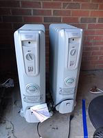 Photo showing two DeLonghi Safeheat space heaters side by side on concrete surface with brick background, featuring front controls and attached safety instructions.