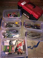 Photo showing a red Craftsman toolbox and five clear plastic bins filled with assorted hand tools including pliers, screwdrivers, wrenches, and miscellaneous hardware items.