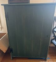 Front view of the vintage green wooden cabinet with door closed showing the vertical paneling, black metal hinges, and wooden knob handle.