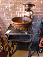Front view of wooden bowl carved into seated Asian figure on wooden table against brick wall.
