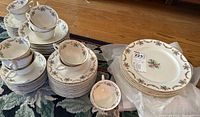 Full view of the entire china set consisting of stacked cups, saucers, plates, and bowls on wood floor with floral carpet.