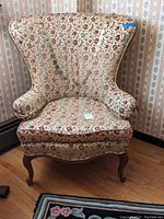 Front view of a vintage wing back chair featuring floral upholstery and signs of wear including stains and arm protectors.