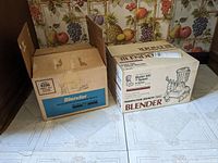 Two Hamilton Beach blender boxes, models 600 and 620G, placed on a floor with fruit-themed wallpaper in background.