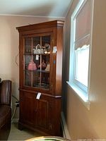 Corner wood-glass door cabinet positioned in corner next to window and seating, showcasing overall form and condition.