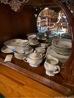 Porcelain dinnerware including plates, cups, saucers, creamer, and sugar bowl stacked on a wooden shelf under a cabinet. Floral designs and gold accents visible.