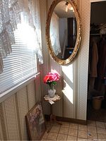 Photo showing corner table under window with glass vase and floral picture on floor leaning against wall, plus ornate oval mirror on adjacent wall.