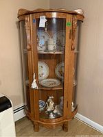 Front angled view of wood display cabinet filled with decorative plates and figurines. Curved glass panels on door and sides. Cabinet door locked with visible key.