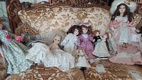 Wide shot of 9 porcelain dolls on vintage sofa showing the assortment including bride and groom dolls, with various formal attire styles and colors.