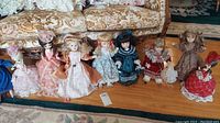 Nine vintage display dolls arranged in a row on a wooden floor in front of a floral sofa; dolls dressed in old-fashioned dresses with various colors and accessories.