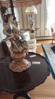 Two porcelain figure lamps with male and female dancing figures sitting on ornate floral bases, each topped with a fringed lamp shade with rips.