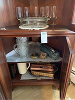 Cabinet interior showing large cut glass pitcher, small coasters, metal serving utensils, pink plastic pitcher, plastic trays, and other kitchenware