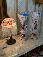 Photo showing two ornate Italian vases with detailed floral and scrollwork designs in pastel colors next to a lamp with a painted floral dome shade and crystal fringe on a metal base.
