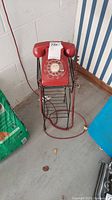 Red rotary dial phone mounted on black metal stand, placed in a corner against a white wall and striped wallpaper. Phone has a coiled red handset cord and visible electrical cord extending from the phone base.