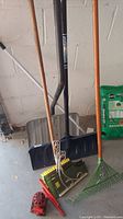 Photo shows two snow shovels with black and metal blades, rake with green plastic tines, yellow broom, and metal hooks on the concrete floor next to a bag of garden soil.