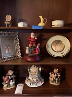 Wide view of shelf with figurines, anniversary plate with floral design, and Santa musical snow globe shown on different shelves.