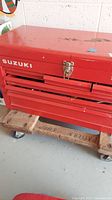 Closed Suzuki red metal tool chest showing wear including paint chipping, scratches, and rust, on a wooden dolly with wheels.