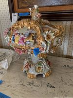 Front side view of ornate porcelain tureen showing detailed hand-painted figures, porcelain floral accents, and gold trim.