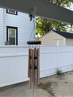 First wind chime hanging under porch roof seen against white fence and house.