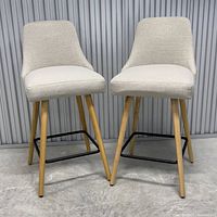 Front view of two beige fabric barstools with wooden legs and black metal footrests on a concrete floor.