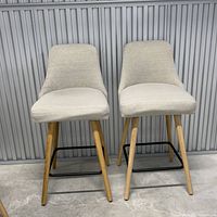 Front view of two beige fabric barstools with wooden legs and black metal footrests against a gray background.