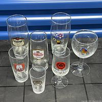 7 varied beer glasses with different brewery logos including G.S., Kirin, Holsten, Saffen, and more, displayed on black grid surface with blue background