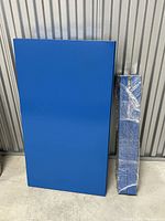 Photo showing blue rectangular table top and separately wrapped legs standing next to each other on concrete floor with metal wall background.