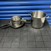 Set of three nested saucepans with black handles and one large stockpot with dual handles shown on black table against blue background.