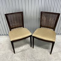 Two wooden dining chairs with cane woven backs and upholstered beige seats, viewed from front against a metal wall.