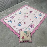 Full image of kids quilt laid out flat on a concrete floor, shows pink scalloped borders and embroidered flower and mushroom designs.