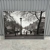 Framed black and white poster showing an Amsterdam canal with a bicycle and streetlamp, photographed standing on floor in front of a striped wall.