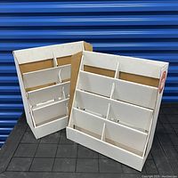 Two white cardboard magazine stands with dividers, shown side by side on a black surface with a blue background. Stands show some darker marks and a TV Guide sticker on one stand.