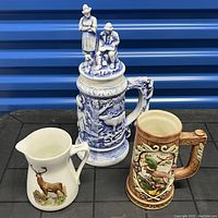 Front view of three items: blue and white stein featuring figurines on lid and wildlife reliefs, cream pitcher with deer painting, brown wood-like beer stein.