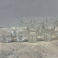 Eight glasses arranged in two rows, mix of faceted and smooth tall clear drinking glasses.