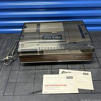 Full view of Zenith Beta Video Director VR9000W with closed transparent dust cover, showing wood-grain front panel, control buttons, power cord, and original documentation.