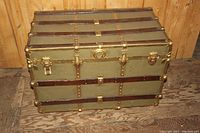 Front angle view of antique trunk showing green metal panels, wood slats, and brass hardware on wooden floor background.