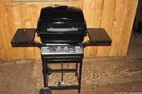 Front view of small black portable gas BBQ grill with two side shelves and control knobs.