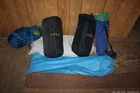 Four camping bags arranged on wooden floor, including two black O.A.S.I.S branded bags, one blue-green bag with black drawstring, and one blue rolled item with cover.