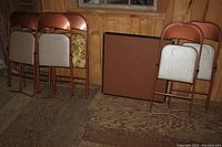 Six metal folding chairs arranged against a wooden wall, three with floral upholstery and three with beige upholstery, with one foldable card table against the wall nearby.