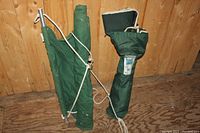 Photo showing folded green camp chair and hammock standing against wooden wall, highlighting carrying bags and white support cords.