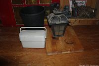 White compost bucket with ventilation slots and handle on top of wooden cutting board beside black planting pot and outdoor lantern light