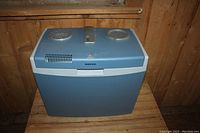 Front view of blue Mobicool plug-in cooler on wooden floor, showing vents, handle, and logo.