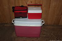 Three coolers placed on floor, showing front and top views. Large Rubbermaid cooler with white lid and handles, medium Igloo cooler with red handle, small red and black soft sided cooler bag.