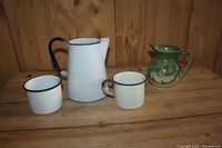 Photo showing four items: a tall white enamelware jug with black handle, two white enamelware mugs with black rims and handles, and a small green ceramic pitcher with a swirl pattern, all placed on a wooden surface against a wood panel background.