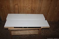 Two white floating shelves placed on a wooden table with a wood-paneled background. Shelves show unfinished particle board undersides and white laminated tops and front edges with slight bevel.