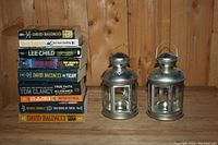 Photo shows two silver metal lanterns and a stack of paperback novels by various thriller authors.