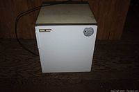Front view of white mini fridge with closed door and power cord lying on the floor.